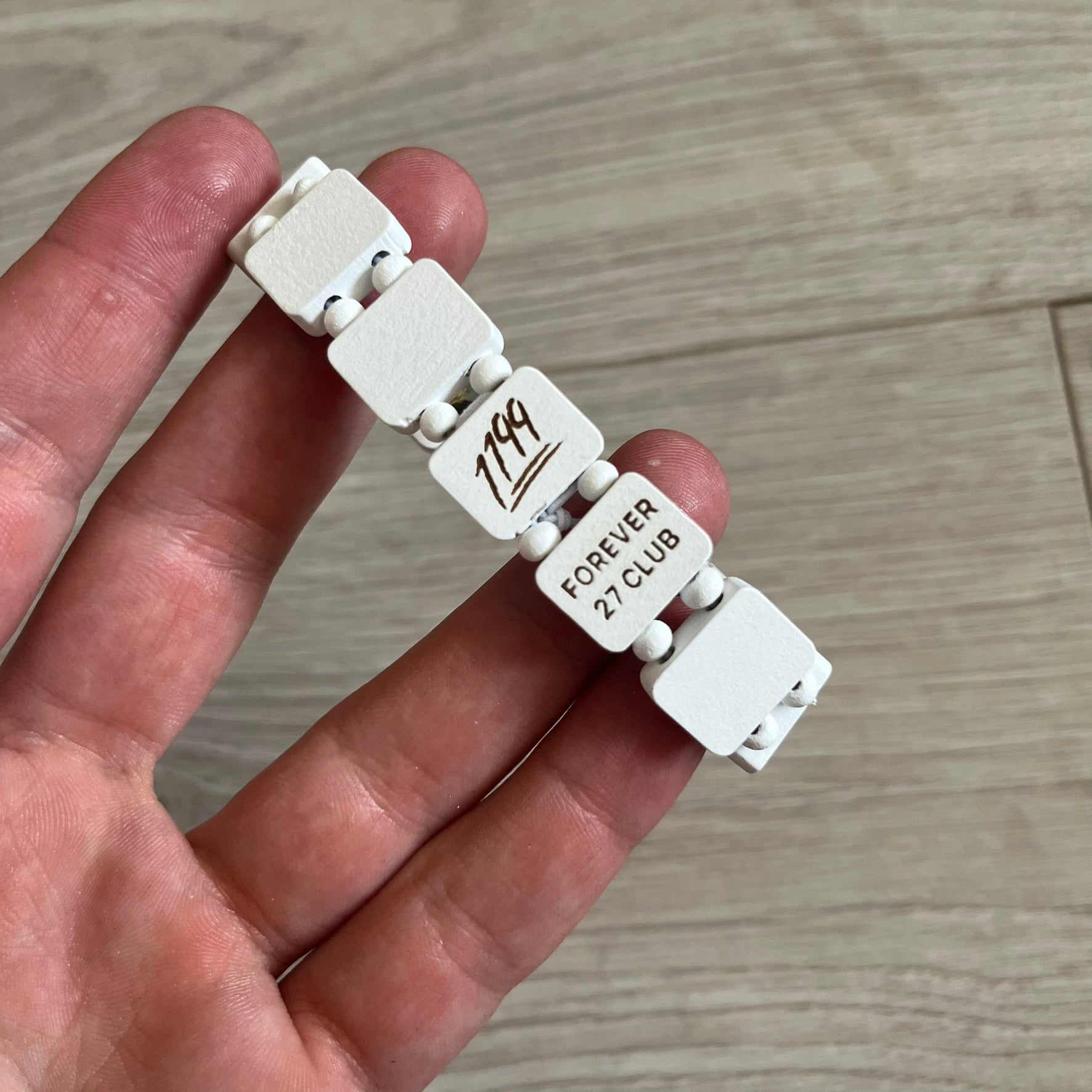 White “Forever 27 Club” bracelet by Bazo 1199, showcasing painted wooden beads and engraved portraits of legendary artists from the infamous 27 Club like Kurt Cobain, Basquiat, and Fredo Santana. Signed “forever 27 club” and “1199” on the inside.