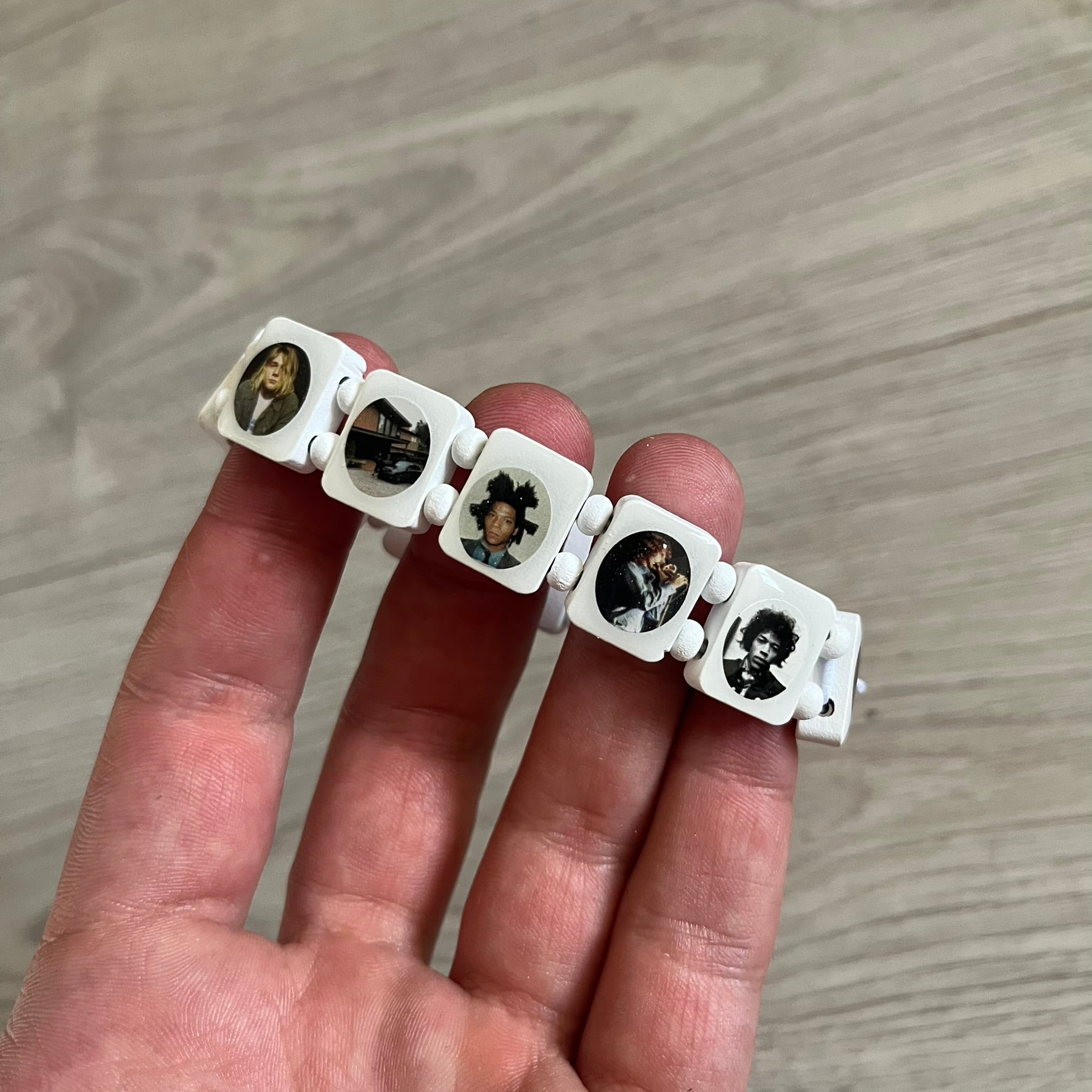 White “Forever 27 Club” bracelet by Bazo 1199, showcasing painted wooden beads and engraved portraits of legendary artists from the infamous 27 Club like Kurt Cobain, Basquiat, and Fredo Santana. Signed “forever 27 club” and “1199” on the inside.