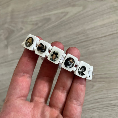 White “Forever 27 Club” bracelet by Bazo 1199, showcasing painted wooden beads and engraved portraits of legendary artists from the infamous 27 Club like Kurt Cobain, Basquiat, and Fredo Santana. Signed “forever 27 club” and “1199” on the inside.