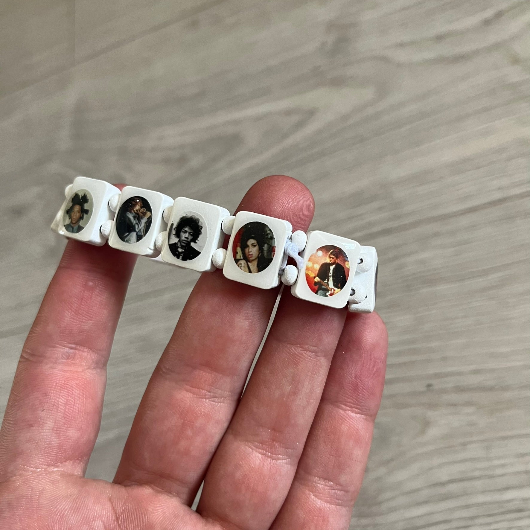 White “Forever 27 Club” bracelet by Bazo 1199, showcasing painted wooden beads and engraved portraits of legendary artists from the infamous 27 Club like Kurt Cobain, Basquiat, and Fredo Santana. Signed “forever 27 club” and “1199” on the inside.