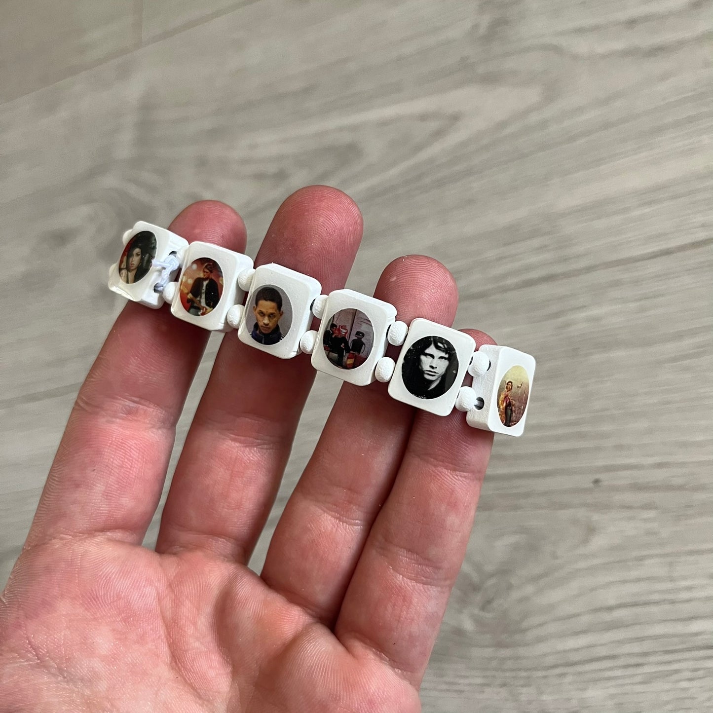 White “Forever 27 Club” bracelet by Bazo 1199, showcasing painted wooden beads and engraved portraits of legendary artists from the infamous 27 Club like Kurt Cobain, Basquiat, and Fredo Santana. Signed “forever 27 club” and “1199” on the inside.