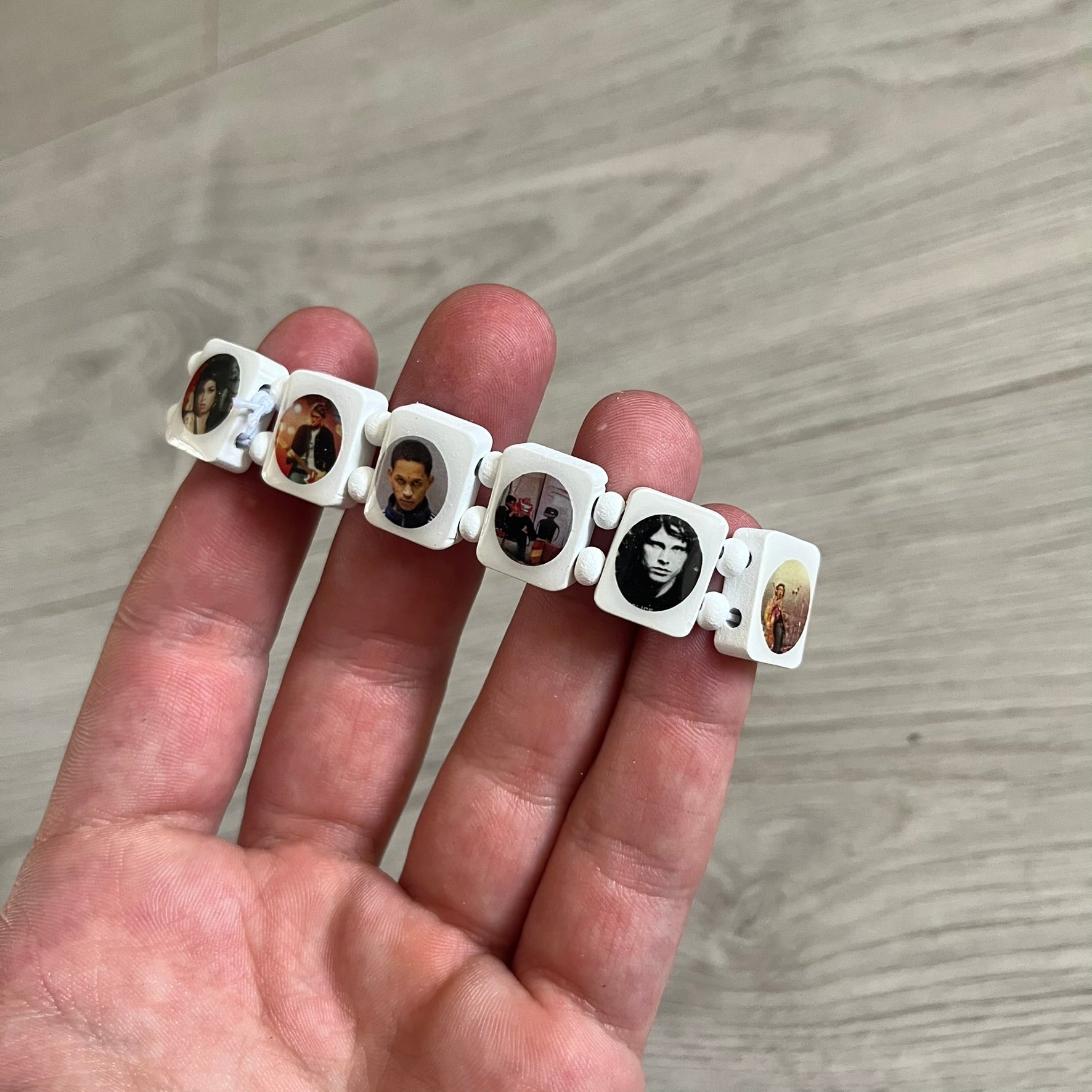White “Forever 27 Club” bracelet by Bazo 1199, showcasing painted wooden beads and engraved portraits of legendary artists from the infamous 27 Club like Kurt Cobain, Basquiat, and Fredo Santana. Signed “forever 27 club” and “1199” on the inside.