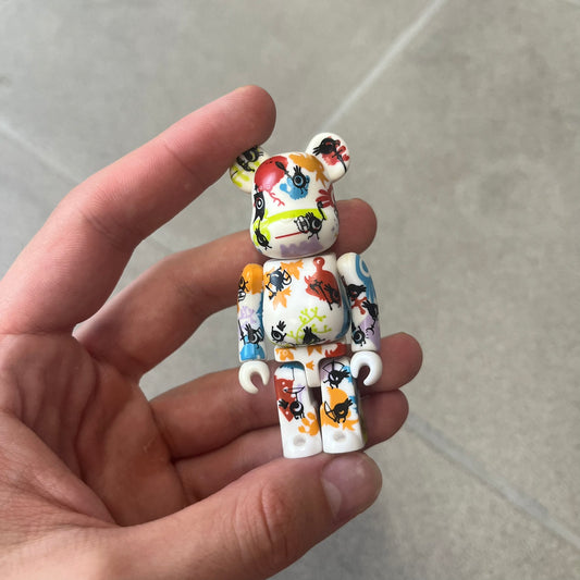 Bearbrick Series 17 Pattern Patapon 2008 Figure features a 100% Bearbrick with a white base covered in multicolored symbols, monsters, and other graphics in red, blue, orange, and green. Black Patapons are layered over the pattern, with a Patapon logo printed on the back. Released in 2008 with a 7.29% rarity, this is an open-box figure with no box or card.
