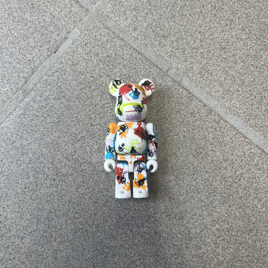 Bearbrick Series 17 Pattern Patapon 2008 Figure features a 100% Bearbrick with a white base covered in multicolored symbols, monsters, and other graphics in red, blue, orange, and green. Black Patapons are layered over the pattern, with a Patapon logo printed on the back. Released in 2008 with a 7.29% rarity, this is an open-box figure with no box or card.