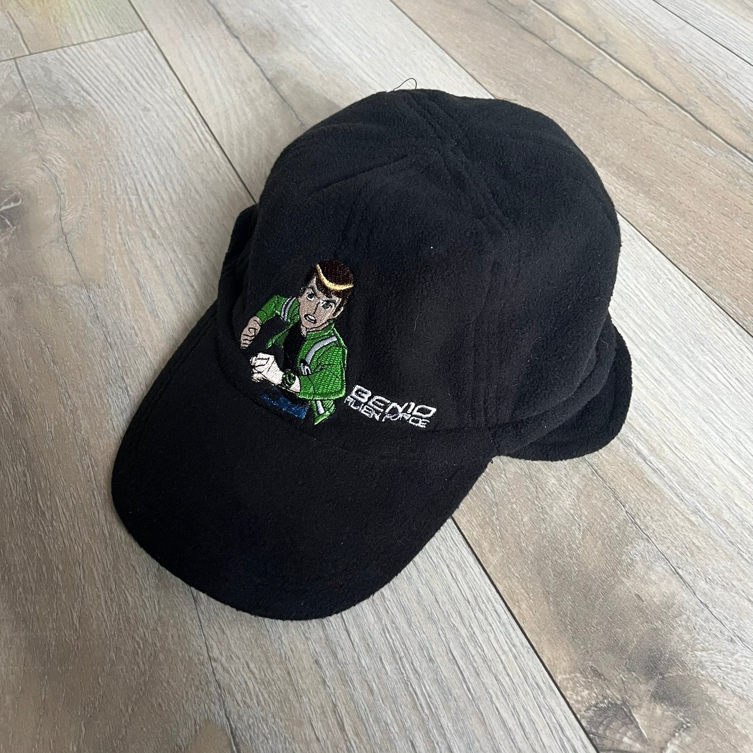 Ben 10 Alien Force Earflap 2011 Hat is a black polyester kids-size hat with green interior lining and earflaps on both sides. The front features embroidered Ben 10 character graphics with “Ben 10 Alien Force” text. Finished with an official 2011 Cartoon Network tag and a soft overall construction.