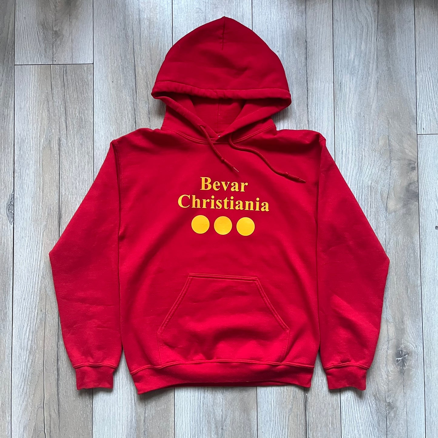 Bevar Christiania Red Hoodie is a red pullover hoodie made on a Gildan Heavy Blend blank, manufactured in Honduras. It features a classic construction with an adjustable hood and a front kangaroo pocket. The front displays yellow “Bevar Christiania” text with three large yellow dots printed underneath.
