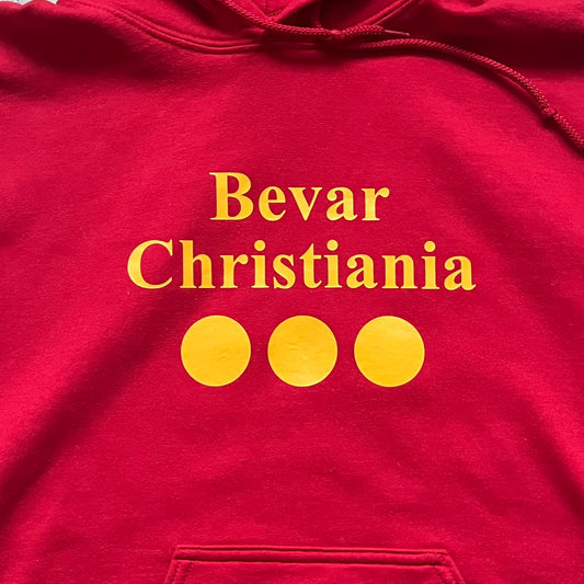 Bevar Christiania Red Hoodie is a red pullover hoodie made on a Gildan Heavy Blend blank, manufactured in Honduras. It features a classic construction with an adjustable hood and a front kangaroo pocket. The front displays yellow “Bevar Christiania” text with three large yellow dots printed underneath.