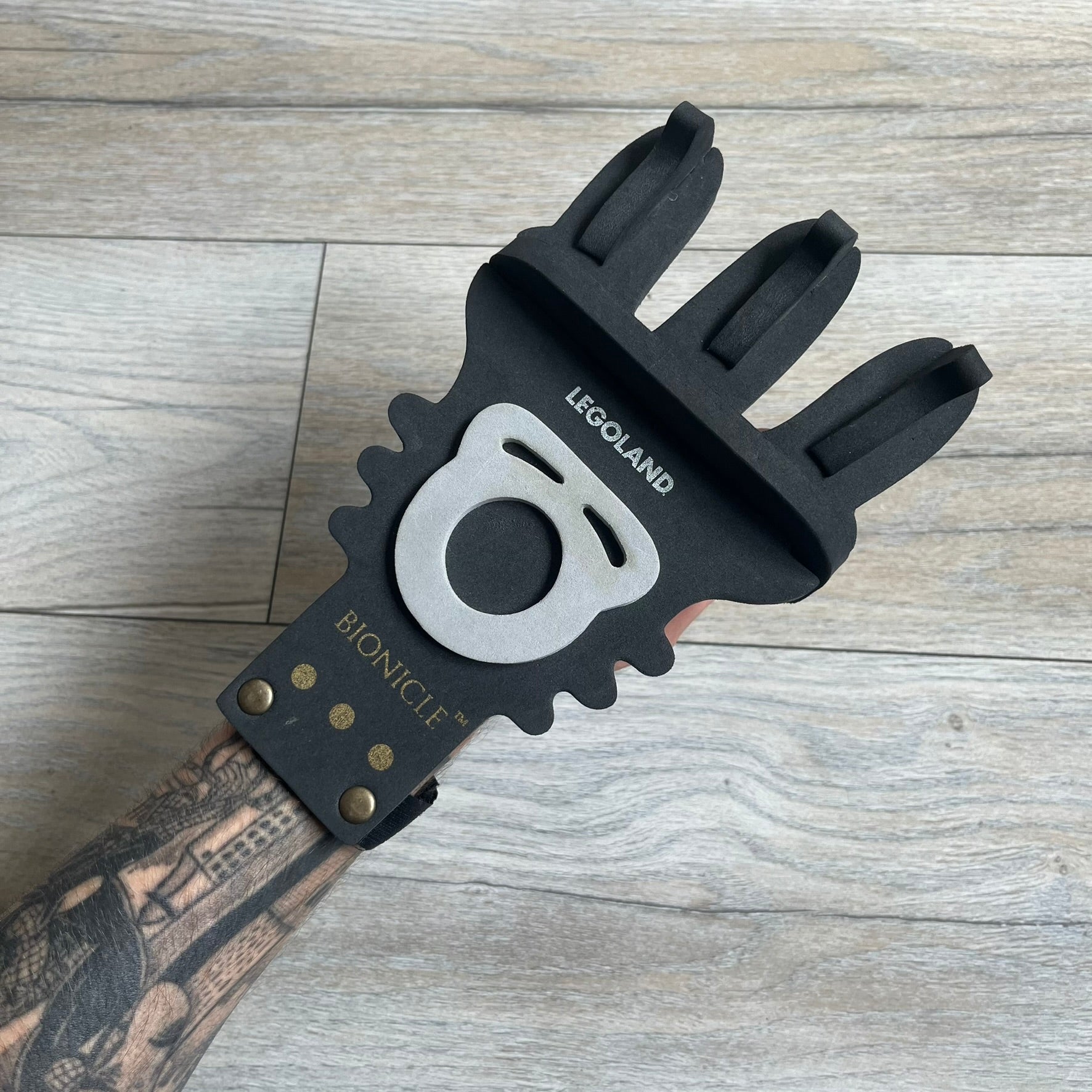 Bionicle 2000s Onua Foam Claw is a black and gray foam weapon featuring three claw tips at the top. It attaches to the wrist with a Velcro strap and is designed to be worn over the hand. The claw is marked with small gray LEGOLAND text and gold Bionicle branding near the bottom.