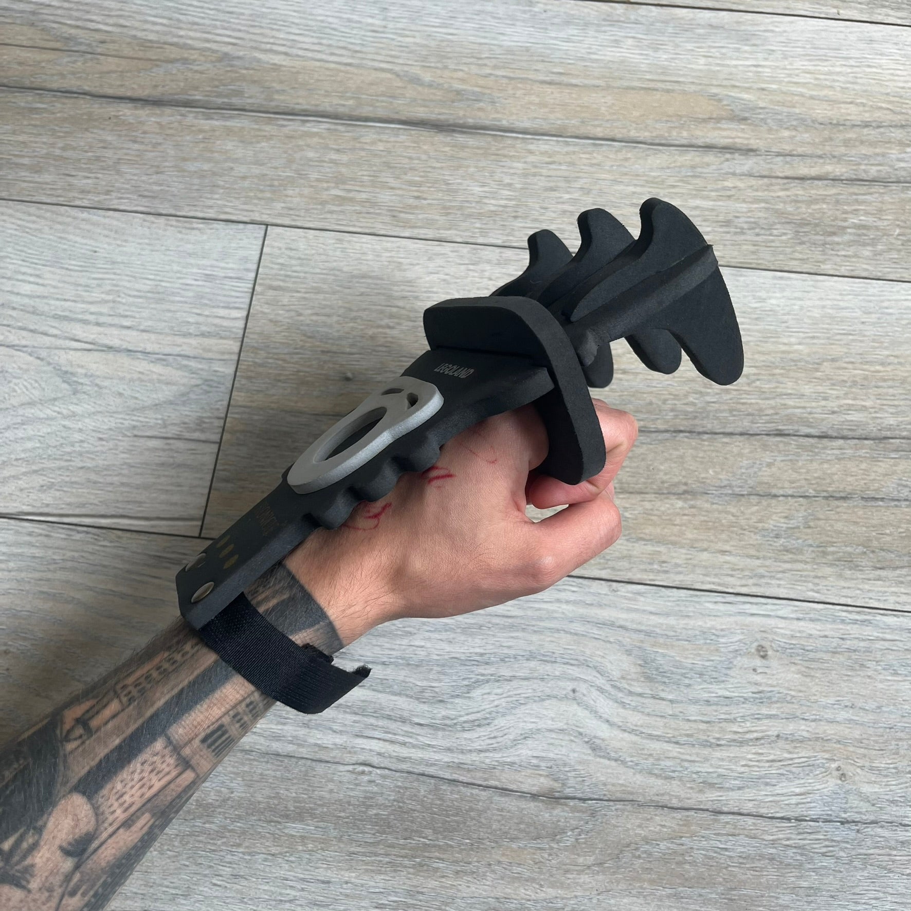 Bionicle 2000s Onua Foam Claw is a black and gray foam weapon featuring three claw tips at the top. It attaches to the wrist with a Velcro strap and is designed to be worn over the hand. The claw is marked with small gray LEGOLAND text and gold Bionicle branding near the bottom.