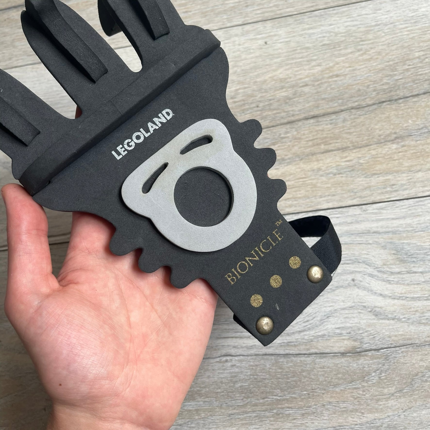 Bionicle 2000s Onua Foam Claw is a black and gray foam weapon featuring three claw tips at the top. It attaches to the wrist with a Velcro strap and is designed to be worn over the hand. The claw is marked with small gray LEGOLAND text and gold Bionicle branding near the bottom.