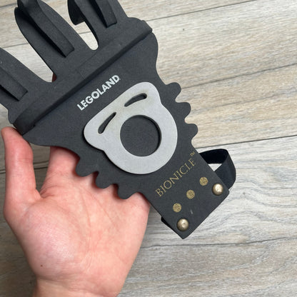 Bionicle 2000s Onua Foam Claw is a black and gray foam weapon featuring three claw tips at the top. It attaches to the wrist with a Velcro strap and is designed to be worn over the hand. The claw is marked with small gray LEGOLAND text and gold Bionicle branding near the bottom.
