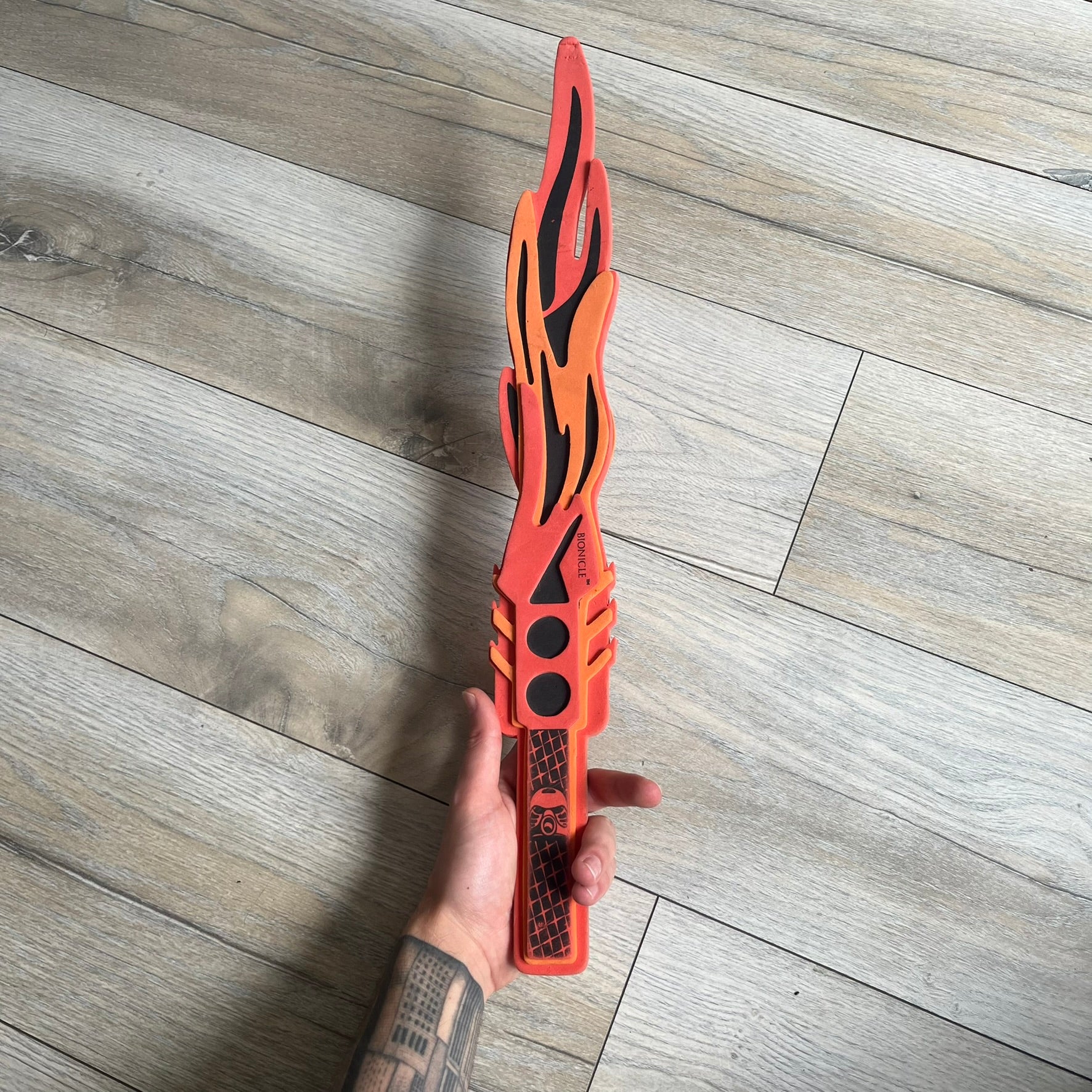 Bionicle 2003 Toa Tahu Flame Sword is a foam toy sword finished in red, orange, and black. The blade features a flame-shaped design at the top, while the handle has a standard grip construction. A small Bionicle mask is attached to the bottom of the handle. The sword is marked with Bionicle and LEGOLAND branding.