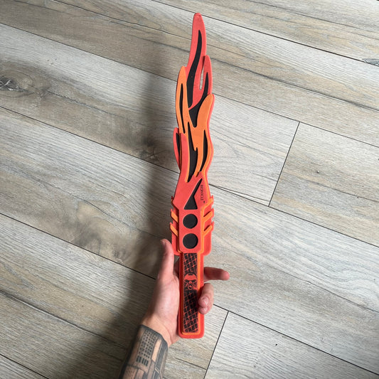 Bionicle 2003 Toa Tahu Flame Sword is a foam toy sword finished in red, orange, and black. The blade features a flame-shaped design at the top, while the handle has a standard grip construction. A small Bionicle mask is attached to the bottom of the handle. The sword is marked with Bionicle and LEGOLAND branding.