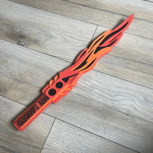 Bionicle 2003 Toa Tahu Flame Sword is a foam toy sword finished in red, orange, and black. The blade features a flame-shaped design at the top, while the handle has a standard grip construction. A small Bionicle mask is attached to the bottom of the handle. The sword is marked with Bionicle and LEGOLAND branding.