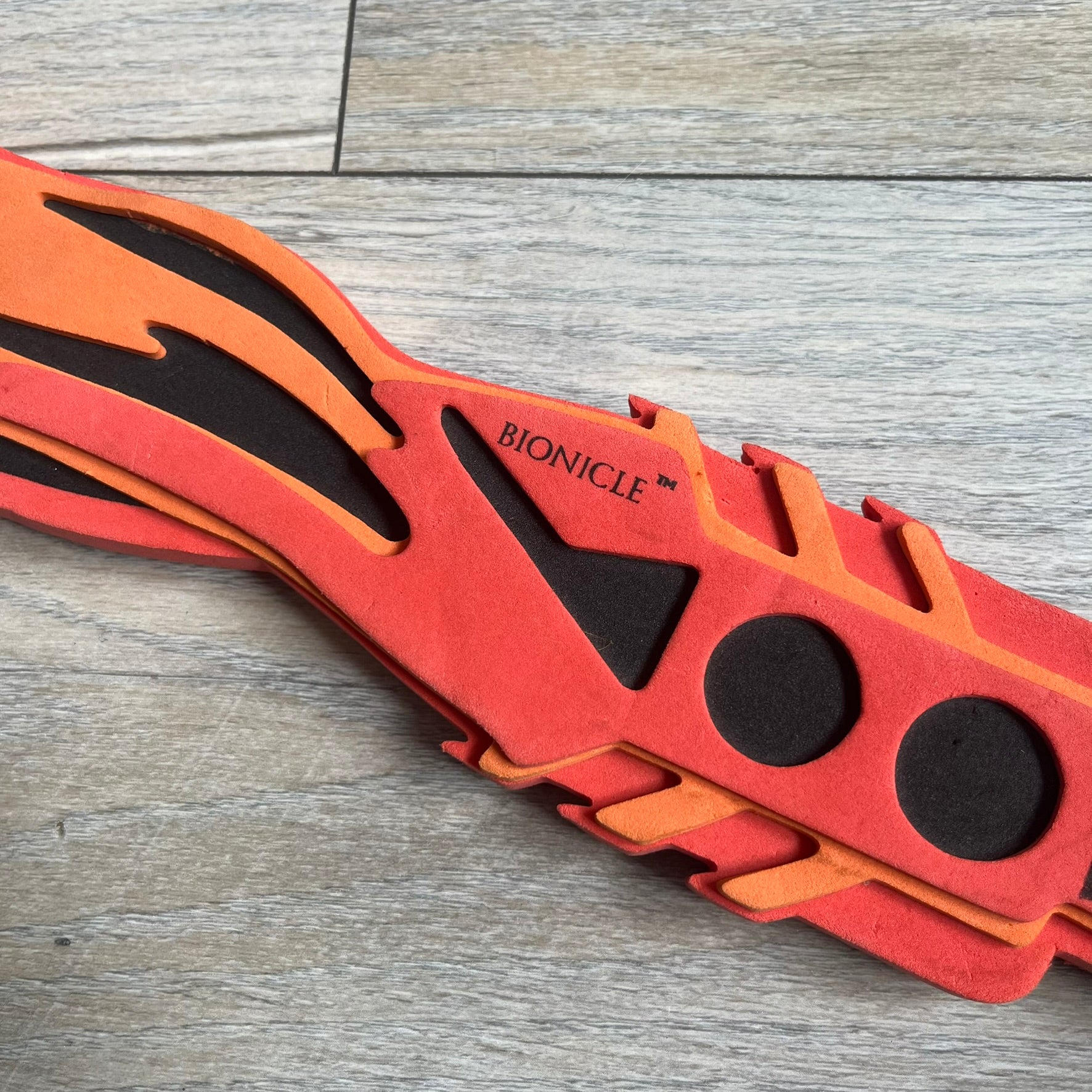 Bionicle 2003 Toa Tahu Flame Sword is a foam toy sword finished in red, orange, and black. The blade features a flame-shaped design at the top, while the handle has a standard grip construction. A small Bionicle mask is attached to the bottom of the handle. The sword is marked with Bionicle and LEGOLAND branding.