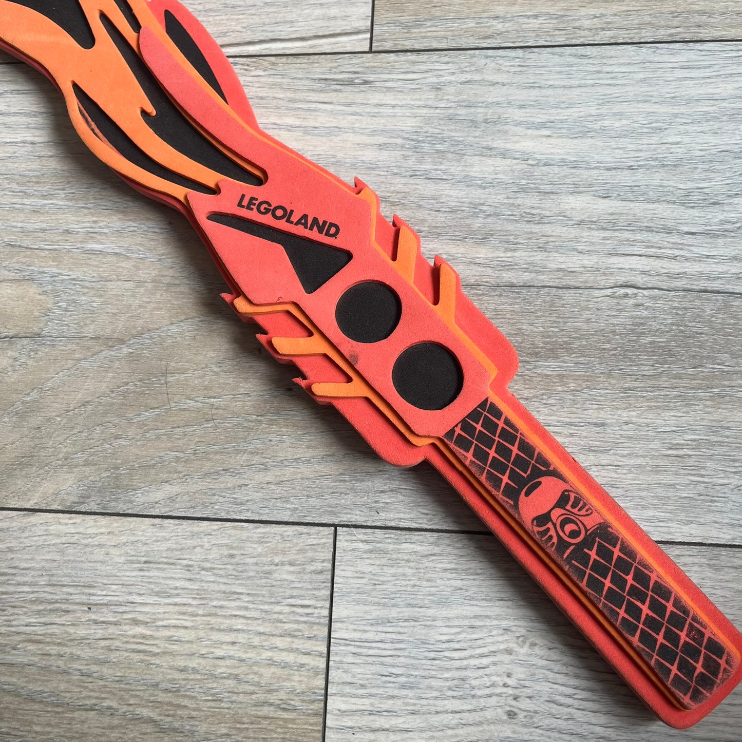 Bionicle 2003 Toa Tahu Flame Sword is a foam toy sword finished in red, orange, and black. The blade features a flame-shaped design at the top, while the handle has a standard grip construction. A small Bionicle mask is attached to the bottom of the handle. The sword is marked with Bionicle and LEGOLAND branding.