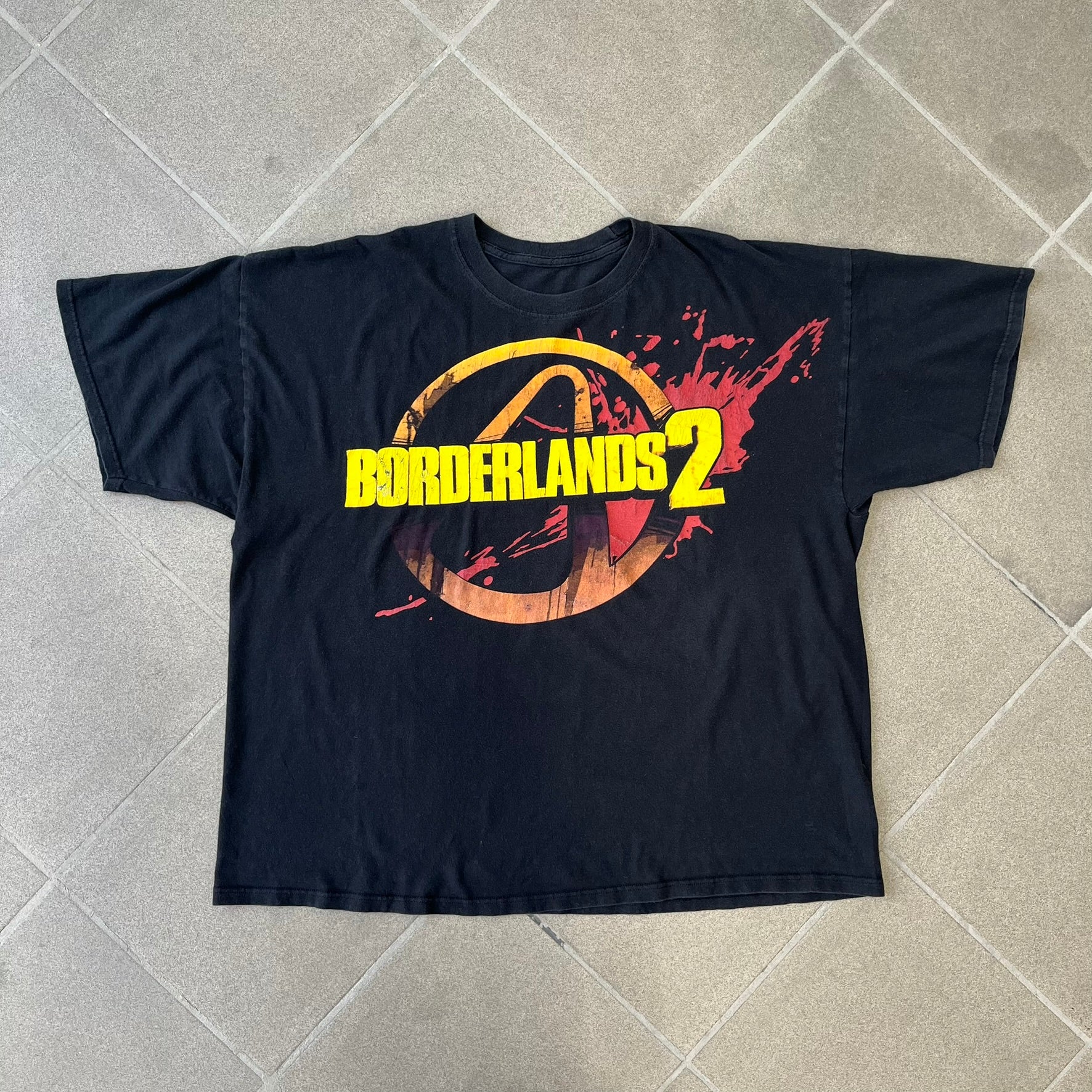 Borderlands 2 Promo 2010's Tee features a black boxy construction with a thick yellow and red Borderlands 2 logo printed on the front. A blood-splatter graphic appears behind the logo, and the shirt is made from 100% cotton.
