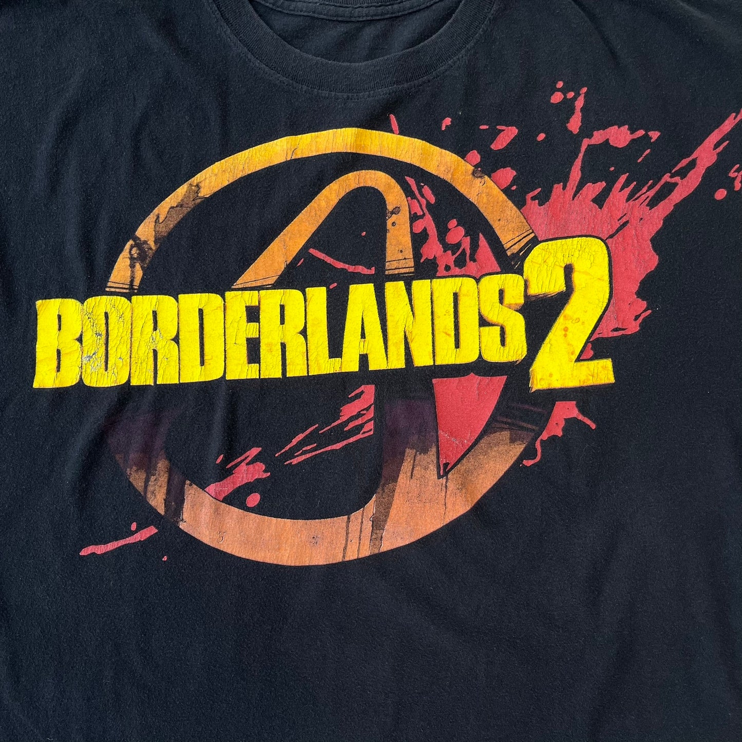 Borderlands 2 Promo 2010's Tee features a black boxy construction with a thick yellow and red Borderlands 2 logo printed on the front. A blood-splatter graphic appears behind the logo, and the shirt is made from 100% cotton.
