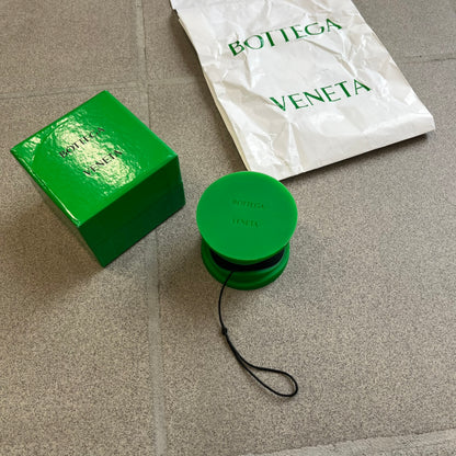 Bottega Veneta Green Rubber Yo-Yo is a collectible accessory crafted from green and black silicone. It features green outer halves with a black center section, a black string, and an engraved Bottega Veneta logo on one side. The yo-yo comes complete with its original box and paper bag.
