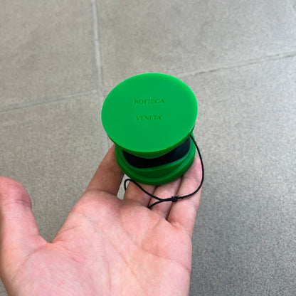 Bottega Veneta Green Rubber Yo-Yo is a collectible accessory crafted from green and black silicone. It features green outer halves with a black center section, a black string, and an engraved Bottega Veneta logo on one side. The yo-yo comes complete with its original box and paper bag.
