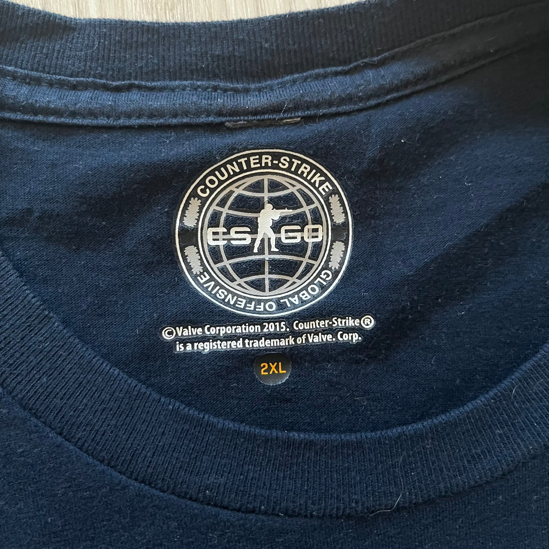 CS:GO 2015 Defuse Kit tee in navy, featuring a bright yellow defuse kit logo on the chest and a small yellow CS:GO patch on the sleeve. Made in Nicaragua from a 60/40 cotton-polyester blend, it includes licensed 2016 Valve Corporation and Counter-Strike Global Offensive tags. A collectible gaming tee that celebrates one of the most iconic in-game items from the series.