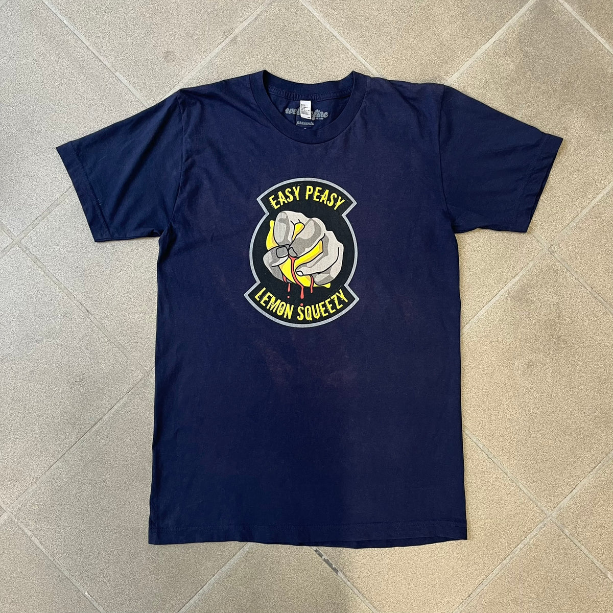 CS:GO Easy Peasy Lemon Squeezy 2015 Tee is a navy T-shirt made in the USA by We Love Fine, featuring a large CS:GO sticker graphic on the front of a hand squeezing a lemon with “Easy Peasy Lemon Squeezy” text. A CS:GO logo is printed on the back below the collar, and it carries a licensed 2015 tag.