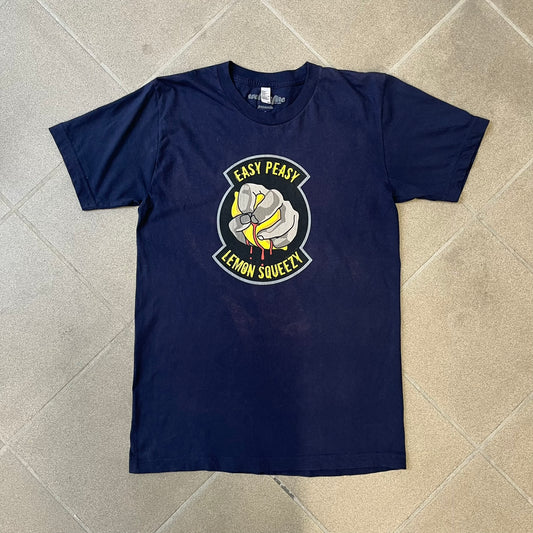 CS:GO Easy Peasy Lemon Squeezy 2015 Tee is a navy T-shirt made in the USA by We Love Fine, featuring a large CS:GO sticker graphic on the front of a hand squeezing a lemon with “Easy Peasy Lemon Squeezy” text. A CS:GO logo is printed on the back below the collar, and it carries a licensed 2015 tag.
