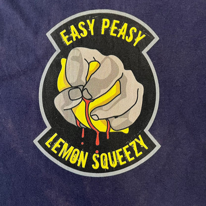 CS:GO Easy Peasy Lemon Squeezy 2015 Tee is a navy T-shirt made in the USA by We Love Fine, featuring a large CS:GO sticker graphic on the front of a hand squeezing a lemon with “Easy Peasy Lemon Squeezy” text. A CS:GO logo is printed on the back below the collar, and it carries a licensed 2015 tag.