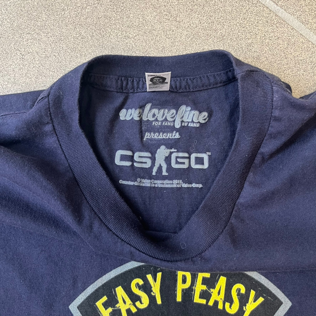 CS:GO Easy Peasy Lemon Squeezy 2015 Tee is a navy T-shirt made in the USA by We Love Fine, featuring a large CS:GO sticker graphic on the front of a hand squeezing a lemon with “Easy Peasy Lemon Squeezy” text. A CS:GO logo is printed on the back below the collar, and it carries a licensed 2015 tag.