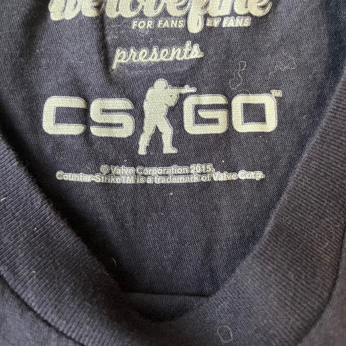 CS:GO Easy Peasy Lemon Squeezy 2015 Tee is a navy T-shirt made in the USA by We Love Fine, featuring a large CS:GO sticker graphic on the front of a hand squeezing a lemon with “Easy Peasy Lemon Squeezy” text. A CS:GO logo is printed on the back below the collar, and it carries a licensed 2015 tag.