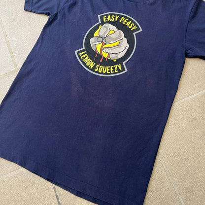 CS:GO Easy Peasy Lemon Squeezy 2015 Tee is a navy T-shirt made in the USA by We Love Fine, featuring a large CS:GO sticker graphic on the front of a hand squeezing a lemon with “Easy Peasy Lemon Squeezy” text. A CS:GO logo is printed on the back below the collar, and it carries a licensed 2015 tag.