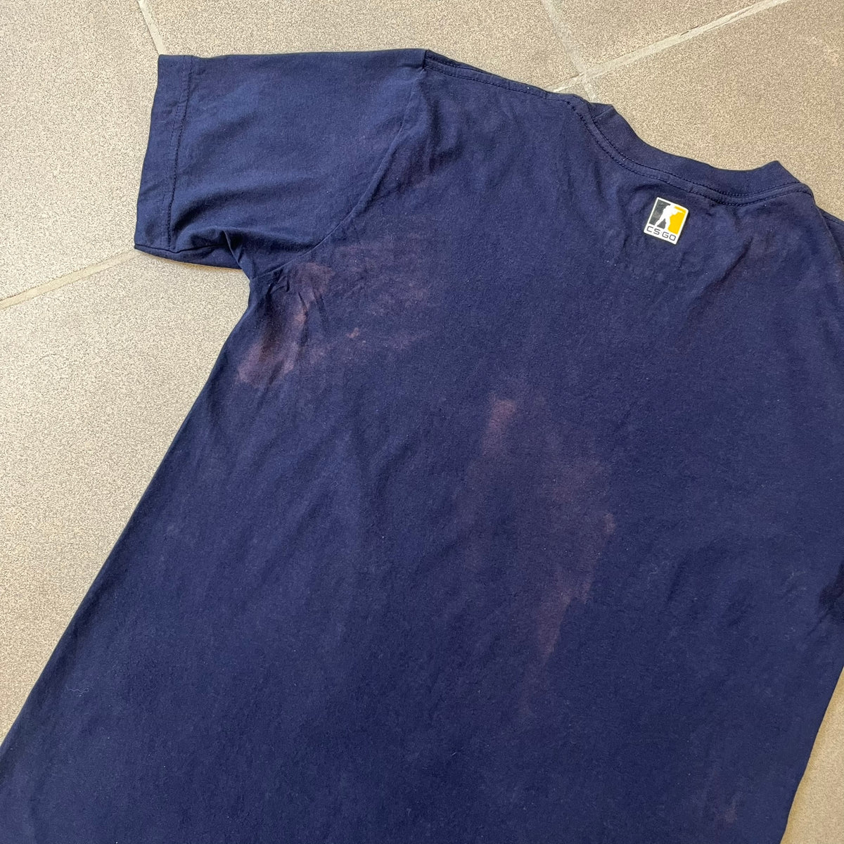 CS:GO Easy Peasy Lemon Squeezy 2015 Tee is a navy T-shirt made in the USA by We Love Fine, featuring a large CS:GO sticker graphic on the front of a hand squeezing a lemon with “Easy Peasy Lemon Squeezy” text. A CS:GO logo is printed on the back below the collar, and it carries a licensed 2015 tag.