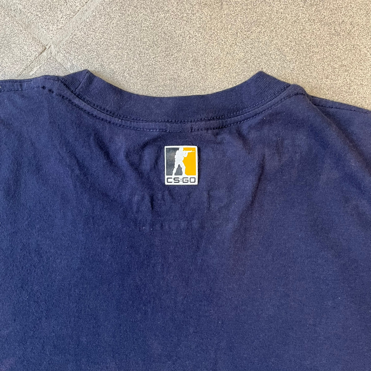 CS:GO Easy Peasy Lemon Squeezy 2015 Tee is a navy T-shirt made in the USA by We Love Fine, featuring a large CS:GO sticker graphic on the front of a hand squeezing a lemon with “Easy Peasy Lemon Squeezy” text. A CS:GO logo is printed on the back below the collar, and it carries a licensed 2015 tag.