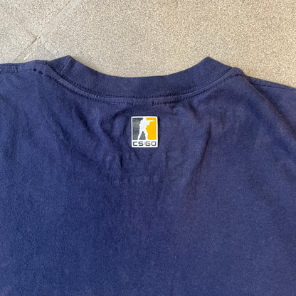 CS:GO Easy Peasy Lemon Squeezy 2015 Tee is a navy T-shirt made in the USA by We Love Fine, featuring a large CS:GO sticker graphic on the front of a hand squeezing a lemon with “Easy Peasy Lemon Squeezy” text. A CS:GO logo is printed on the back below the collar, and it carries a licensed 2015 tag.