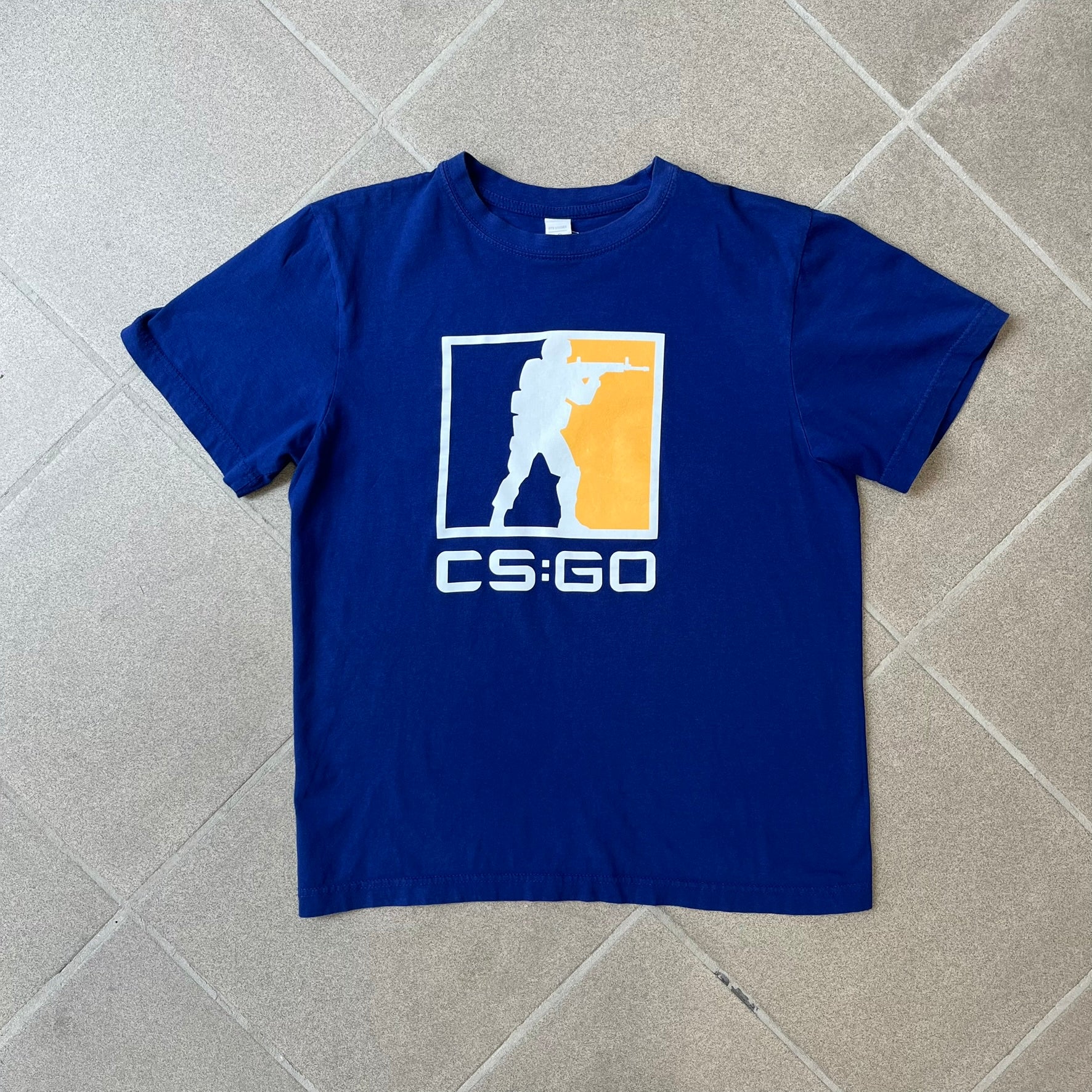CS:GO Logo Navy Tee features a navy blue design with a large Counter-Strike: Global Offensive logo printed across the front. The graphic showcases the iconic navy, orange, and white CS:GO logo with the Counter-Terrorists silhouette above big white "CS:GO" text. Made from 100% cotton.