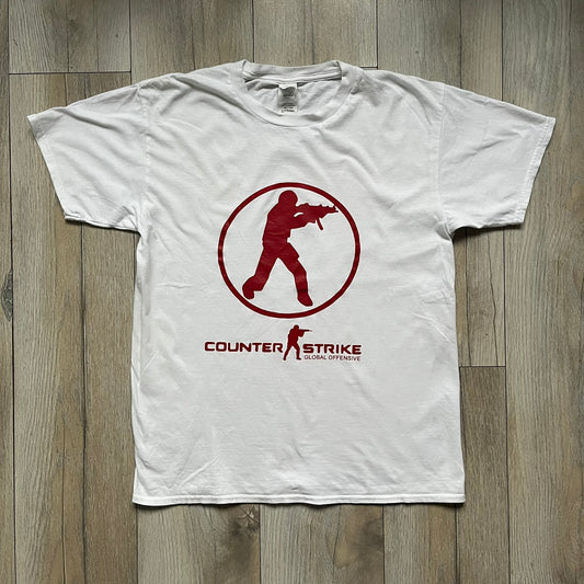 CS:GO White & Red Logo 2010’s Tee is a white T-shirt made from 100% cotton on a Gildan blank. The front features a large red CS:GO logo with an armed figure inside a circle and “Counter-Strike Global Offensive” text below. The shirt reflects early 2010s CS:GO merchandise design.