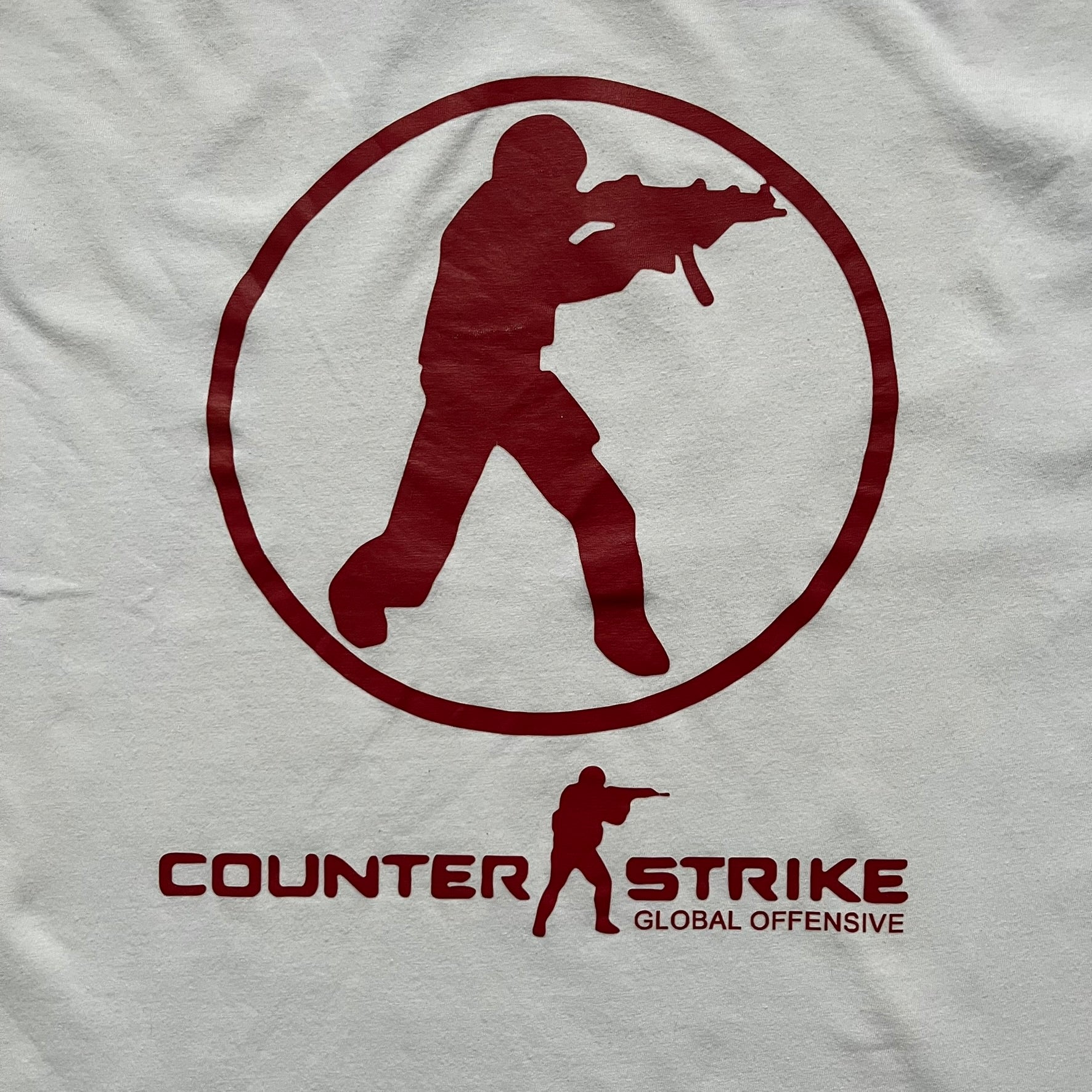CS:GO White & Red Logo 2010’s Tee is a white T-shirt made from 100% cotton on a Gildan blank. The front features a large red CS:GO logo with an armed figure inside a circle and “Counter-Strike Global Offensive” text below. The shirt reflects early 2010s CS:GO merchandise design.