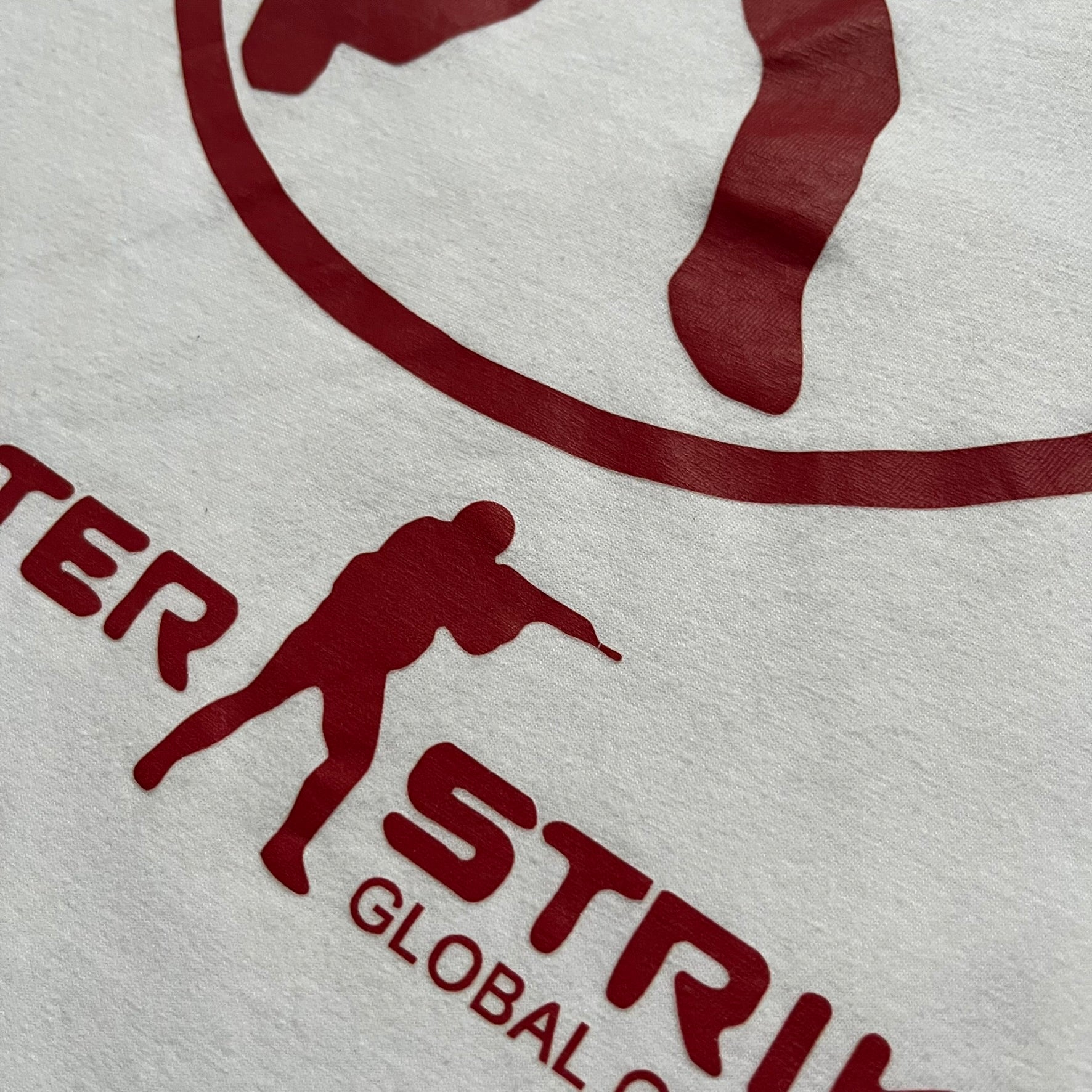 CS:GO White & Red Logo 2010’s Tee is a white T-shirt made from 100% cotton on a Gildan blank. The front features a large red CS:GO logo with an armed figure inside a circle and “Counter-Strike Global Offensive” text below. The shirt reflects early 2010s CS:GO merchandise design.