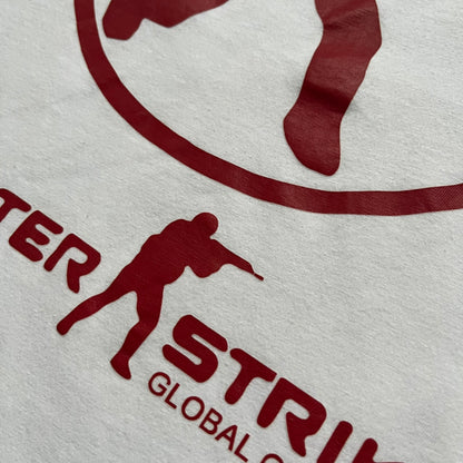 CS:GO White & Red Logo 2010’s Tee is a white T-shirt made from 100% cotton on a Gildan blank. The front features a large red CS:GO logo with an armed figure inside a circle and “Counter-Strike Global Offensive” text below. The shirt reflects early 2010s CS:GO merchandise design.
