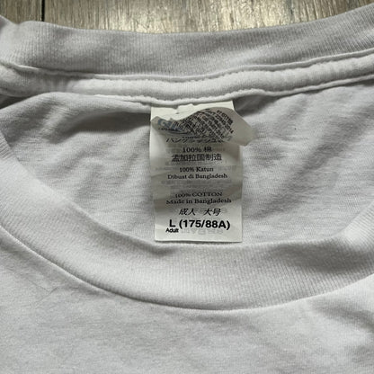 CS:GO White & Red Logo 2010’s Tee is a white T-shirt made from 100% cotton on a Gildan blank. The front features a large red CS:GO logo with an armed figure inside a circle and “Counter-Strike Global Offensive” text below. The shirt reflects early 2010s CS:GO merchandise design.