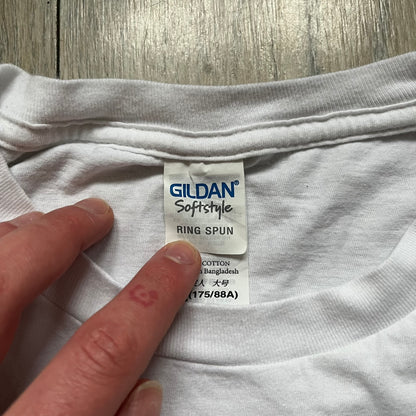 CS:GO White & Red Logo 2010’s Tee is a white T-shirt made from 100% cotton on a Gildan blank. The front features a large red CS:GO logo with an armed figure inside a circle and “Counter-Strike Global Offensive” text below. The shirt reflects early 2010s CS:GO merchandise design.