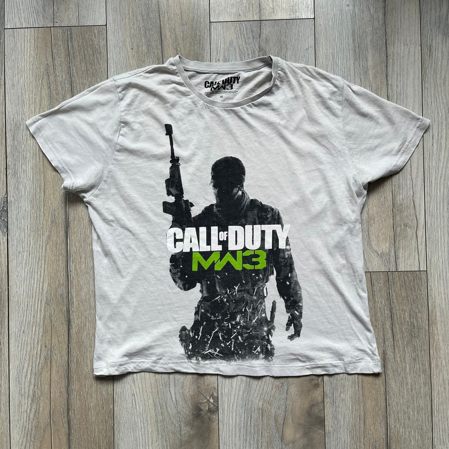 Call of Duty: MW3 2011 Cover Tee is a white to very light beige T-shirt featuring the Call of Duty: Modern Warfare 3 cover artwork printed on the front. The graphic includes the Call of Duty MW3 logo and the game's main cover image in black and gray tones. The shirt includes MW3 licensing on the neck tag.