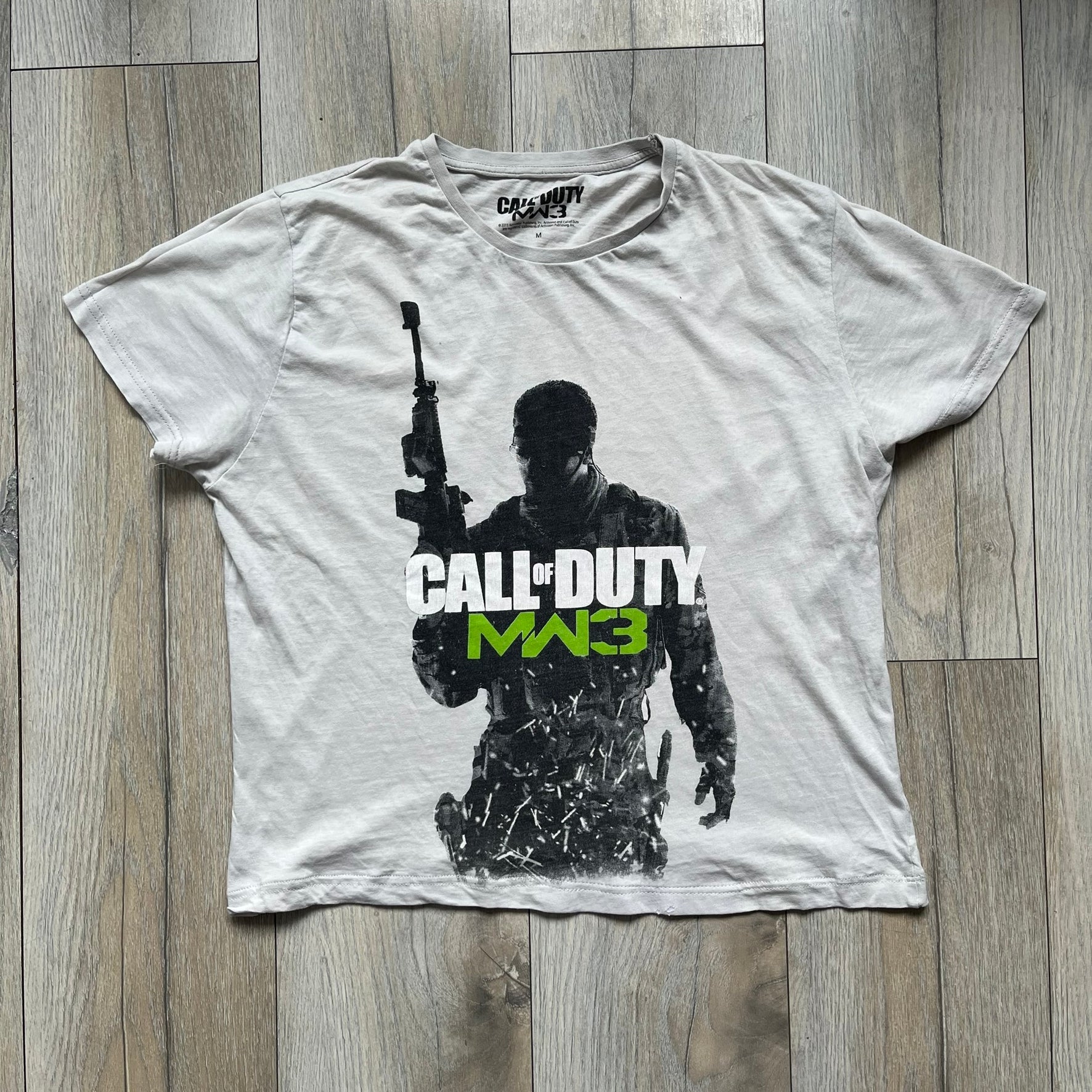 Call of Duty: MW3 2011 Cover Tee is a white to very light beige T-shirt featuring the Call of Duty: Modern Warfare 3 cover artwork printed on the front. The graphic includes the Call of Duty MW3 logo and the game's main cover image in black and gray tones. The shirt includes MW3 licensing on the neck tag.
