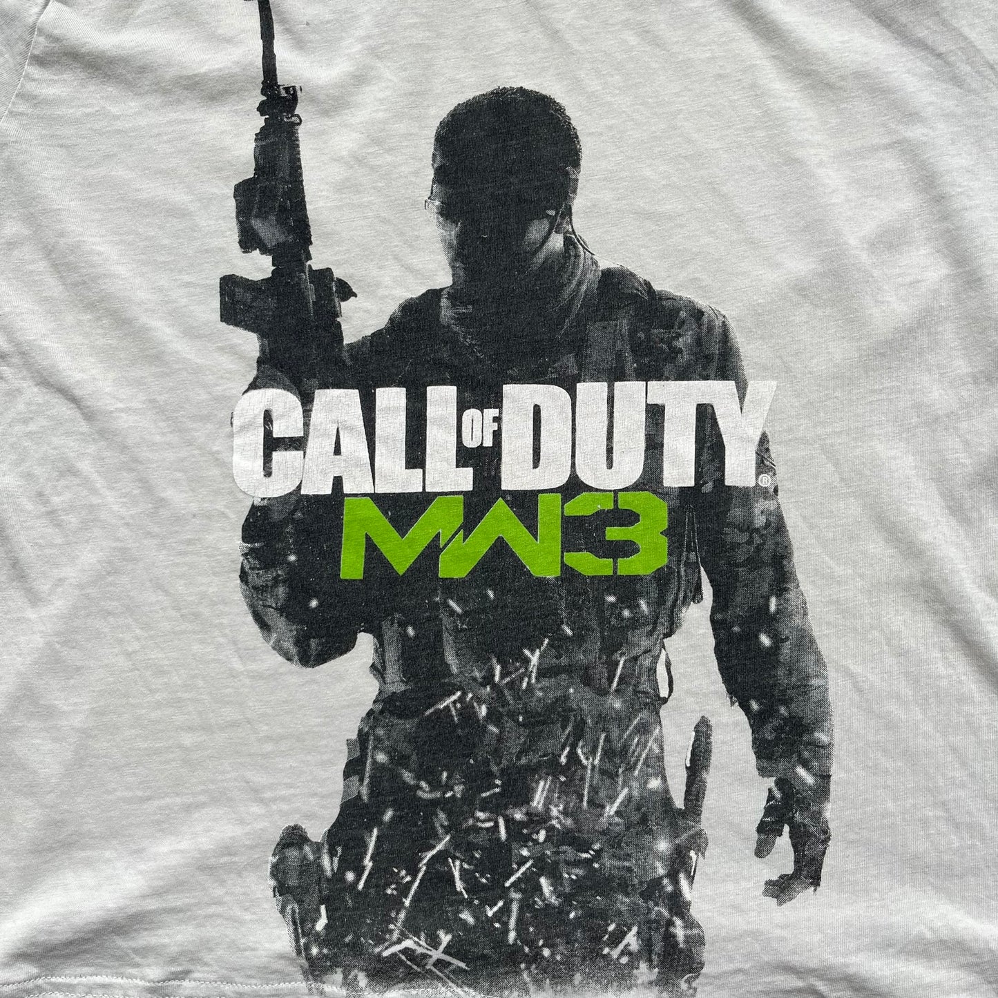 Call of Duty: MW3 2011 Cover Tee is a white to very light beige T-shirt featuring the Call of Duty: Modern Warfare 3 cover artwork printed on the front. The graphic includes the Call of Duty MW3 logo and the game's main cover image in black and gray tones. The shirt includes MW3 licensing on the neck tag.