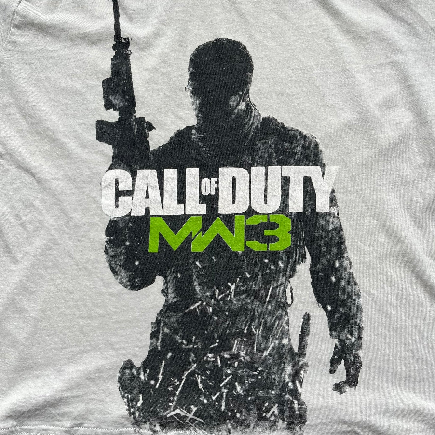 Call of Duty: MW3 2011 Cover Tee is a white to very light beige T-shirt featuring the Call of Duty: Modern Warfare 3 cover artwork printed on the front. The graphic includes the Call of Duty MW3 logo and the game's main cover image in black and gray tones. The shirt includes MW3 licensing on the neck tag.