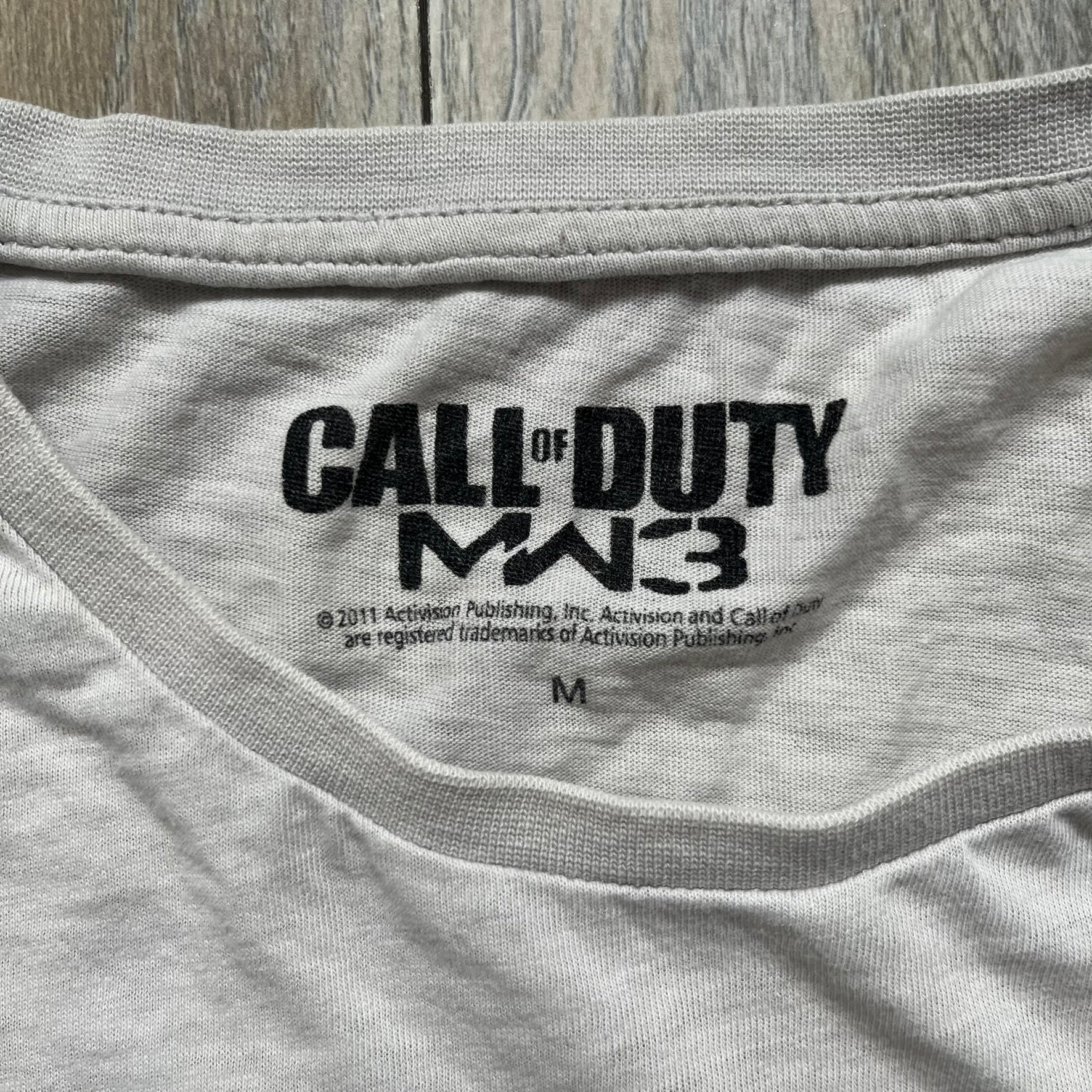 Call of Duty: MW3 2011 Cover Tee is a white to very light beige T-shirt featuring the Call of Duty: Modern Warfare 3 cover artwork printed on the front. The graphic includes the Call of Duty MW3 logo and the game's main cover image in black and gray tones. The shirt includes MW3 licensing on the neck tag.