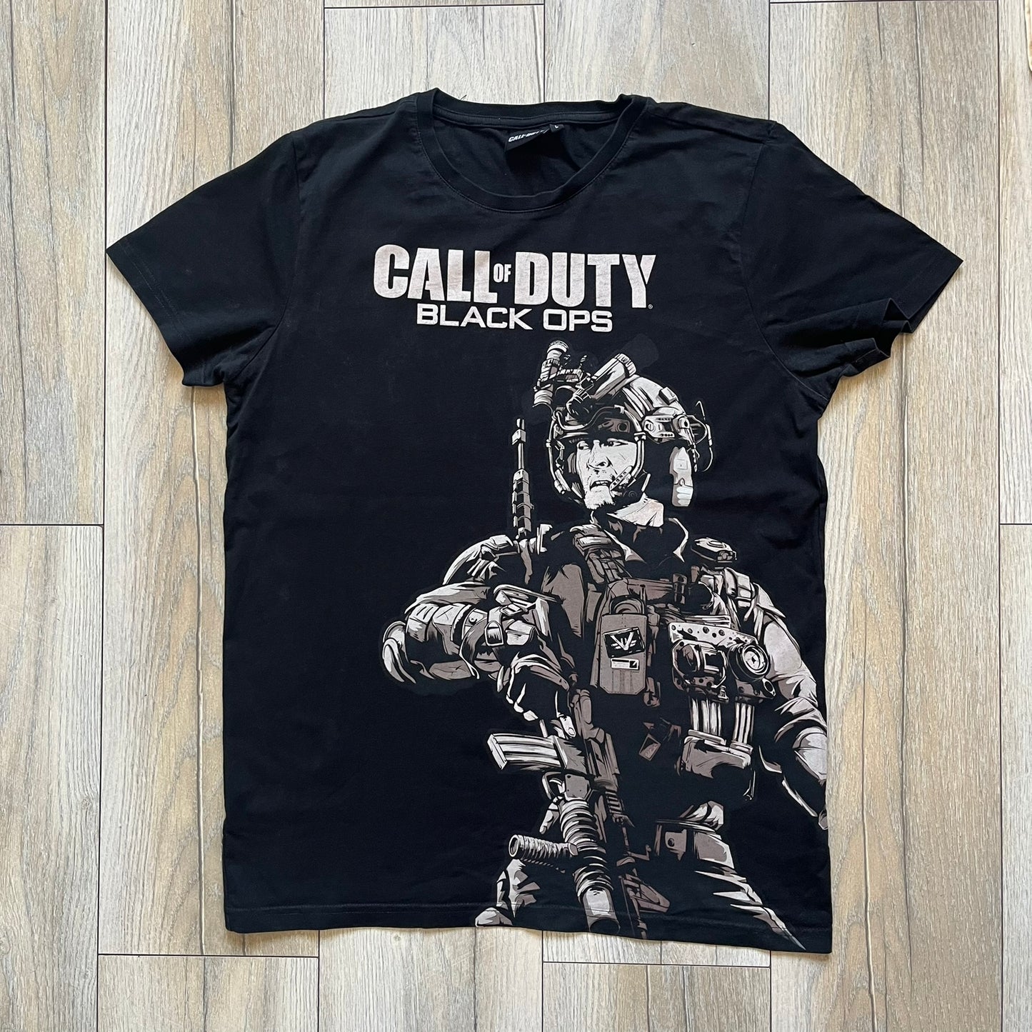 Call of Duty: Black Ops 2018 Black Tee is a black promotional T-shirt with a 2018 Activision Publishing licensed neck tag. The front features a large white and grey character graphic in armor holding a weapon, with the “Call of Duty Black Ops” logo printed above. The print spans most of the front from top to bottom.
