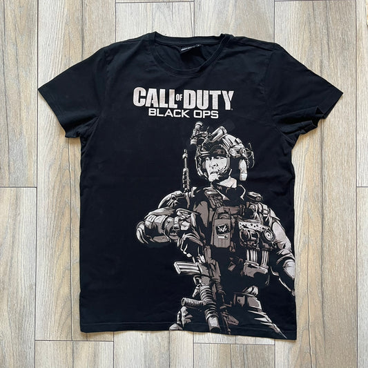 Call of Duty: Black Ops 2018 Black Tee is a black promotional T-shirt with a 2018 Activision Publishing licensed neck tag. The front features a large white and grey character graphic in armor holding a weapon, with the “Call of Duty Black Ops” logo printed above. The print spans most of the front from top to bottom.
