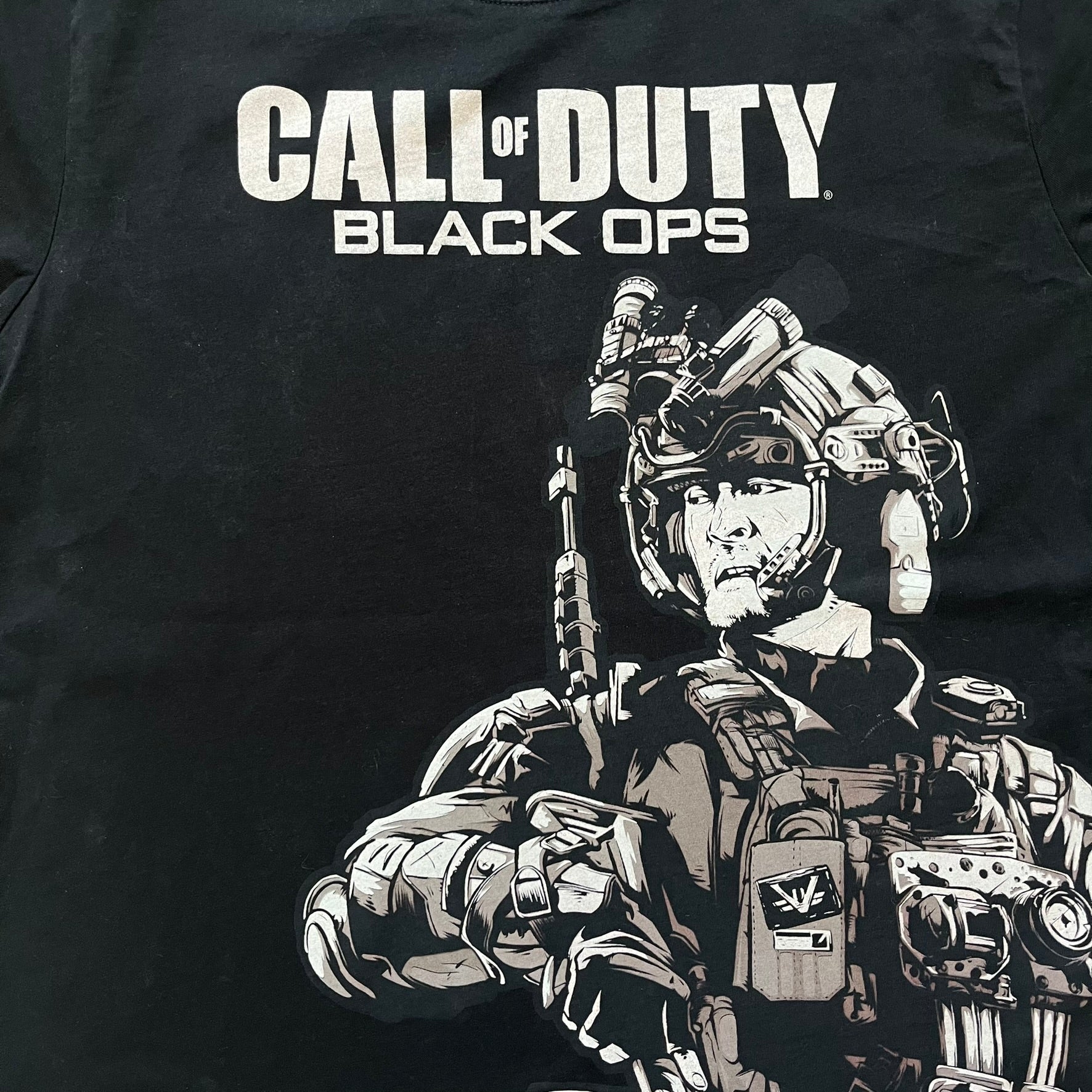 Call of Duty: Black Ops 2018 Black Tee is a black promotional T-shirt with a 2018 Activision Publishing licensed neck tag. The front features a large white and grey character graphic in armor holding a weapon, with the “Call of Duty Black Ops” logo printed above. The print spans most of the front from top to bottom.
