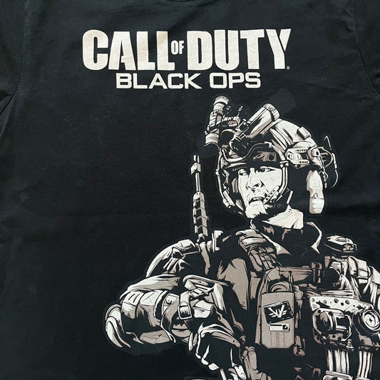 Call of Duty: Black Ops 2018 Black Tee is a black promotional T-shirt with a 2018 Activision Publishing licensed neck tag. The front features a large white and grey character graphic in armor holding a weapon, with the “Call of Duty Black Ops” logo printed above. The print spans most of the front from top to bottom.
